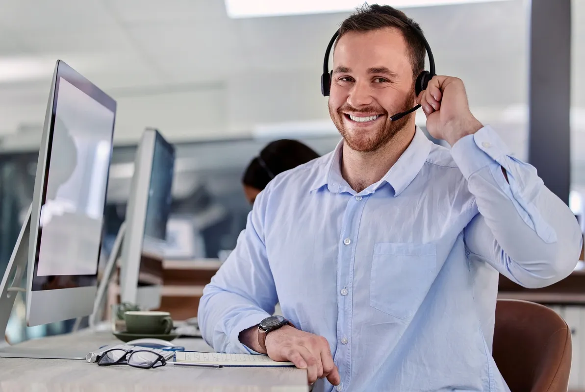 Computer Repair Store Dispatcher With Headset Smiling At Camera In Front Of a Mac Computer